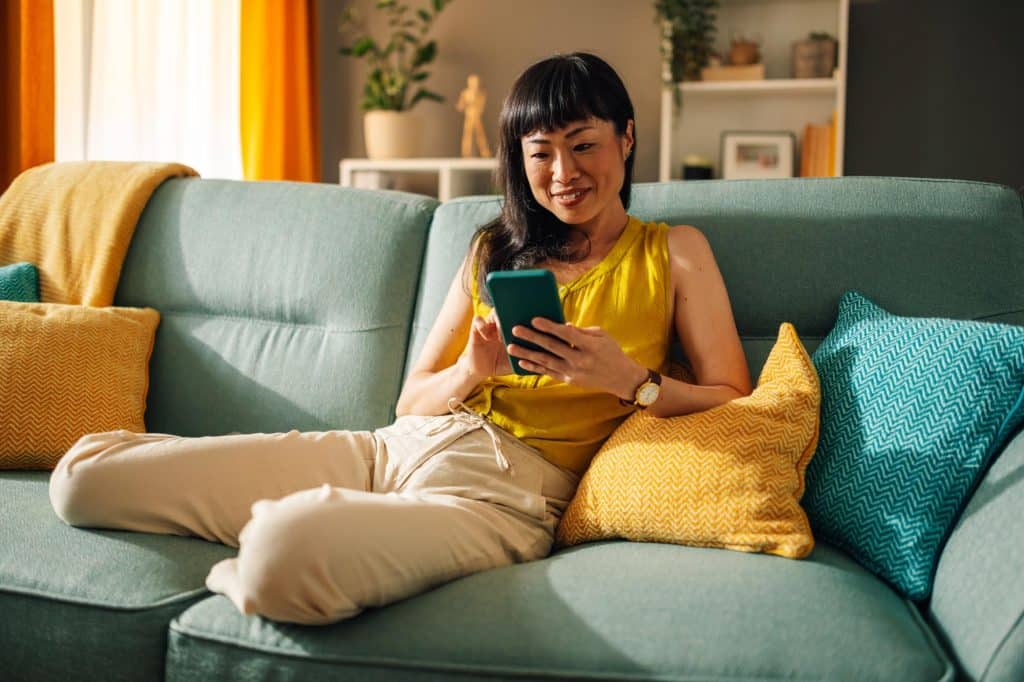 Asian woman enjoying her smartphone on green couch, colorful living