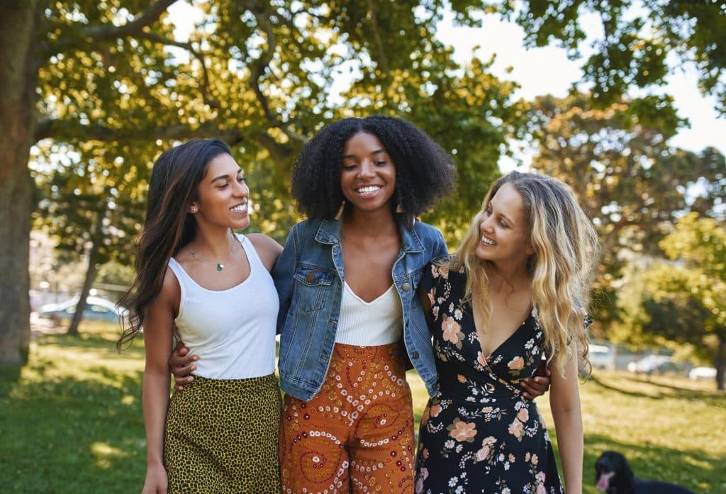 Portrait of a group of happy three diverse young women hugging laughing and having fun in the park on a sunny day while walking