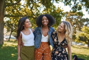 Portrait of a group of happy three diverse young women hugging laughing and having fun in the park on a sunny day while walking